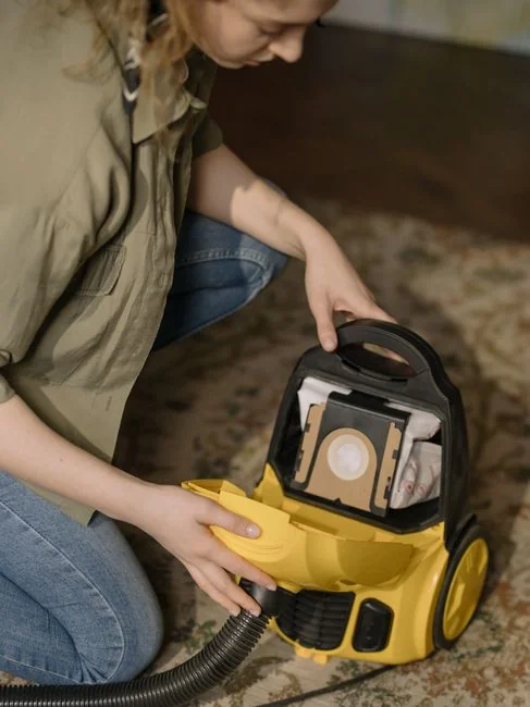 Individual replacing a bag in a yellow vacuum cleaner on a patterned carpet.